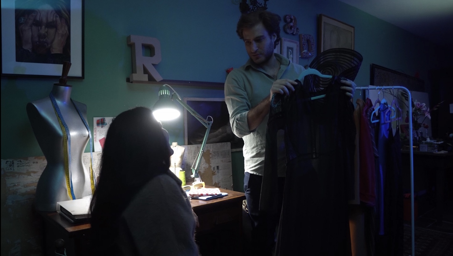Homem segurando um vestido em um cabide enquanto conversa com uma mulher sentada em um ambiente de costura com iluminação suave. Há um manequim com fita métrica, uma luminária acesa sobre a mesa, e roupas penduradas em um suporte ao fundo.